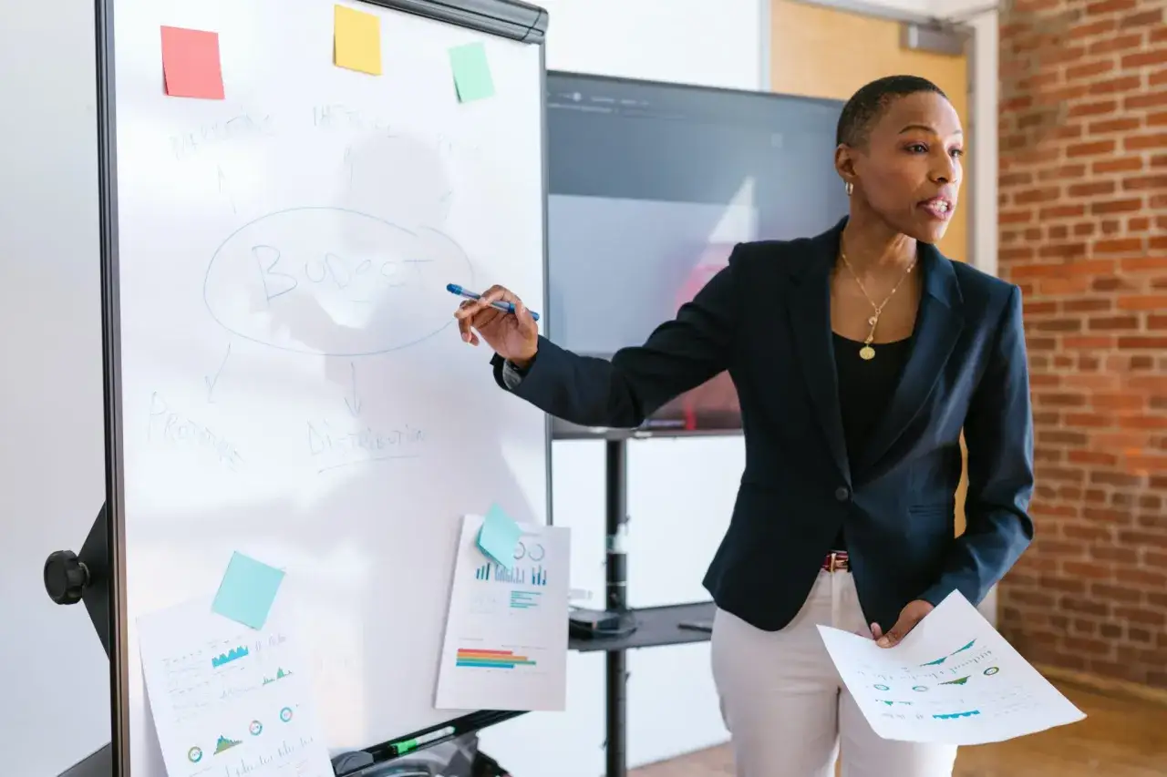 A business coach explains how to find investors for a business, pointing to a whiteboard with a budget diagram.