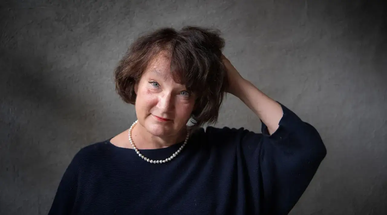 Monika Fagerholm, med en pärlhalsband och mörk tröja, ler mot betraktaren med handen i håret.