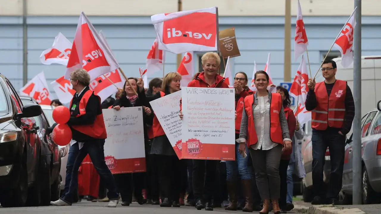 Demonstranten mit roten Westen und Fahnen marschieren. Eine Frau fragt: