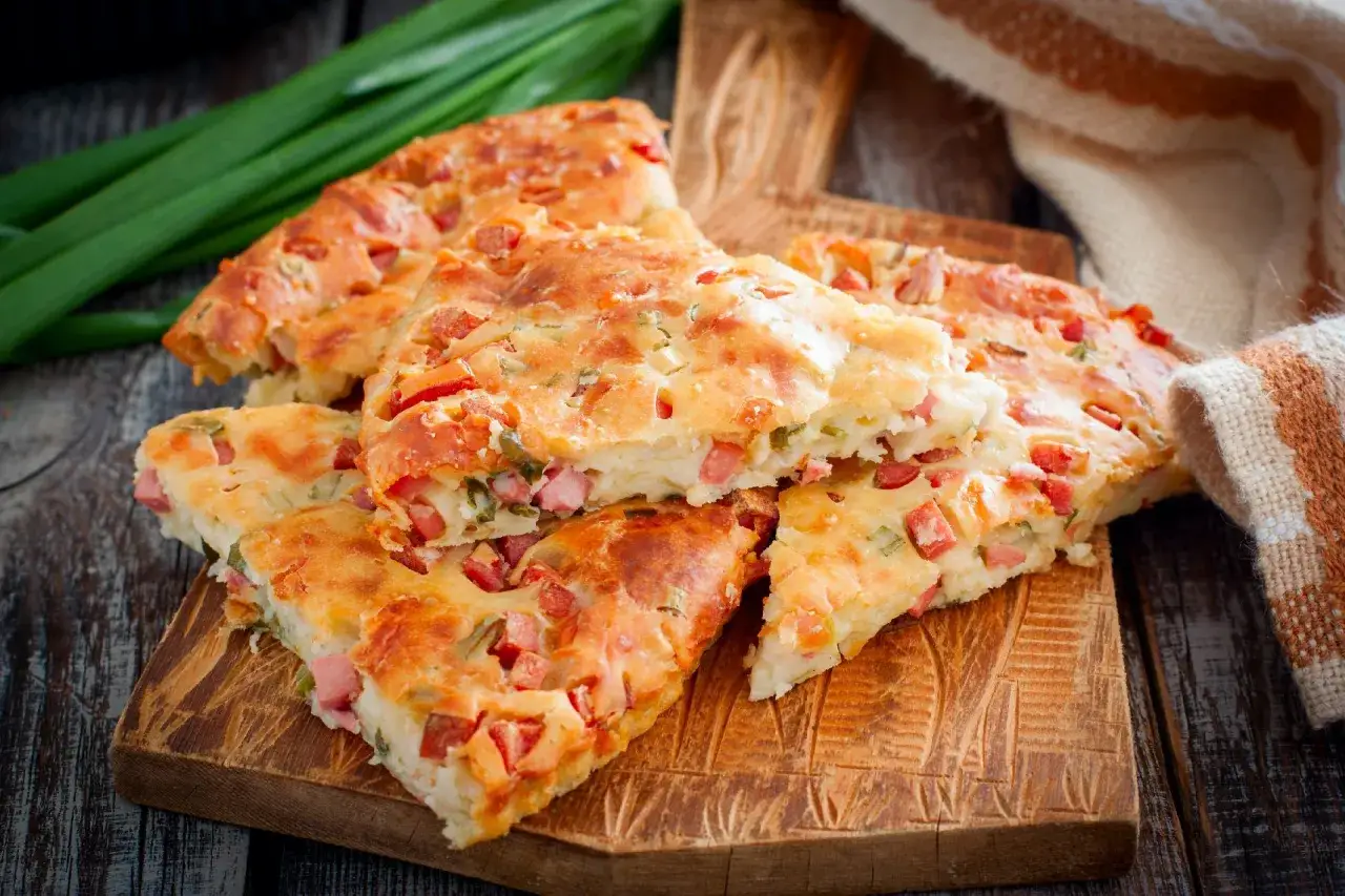 Pyszna tarta z kiełbasą, pokrojona na kawałki, leży na drewnianej desce. Obok zielona cebulka i ręcznik.