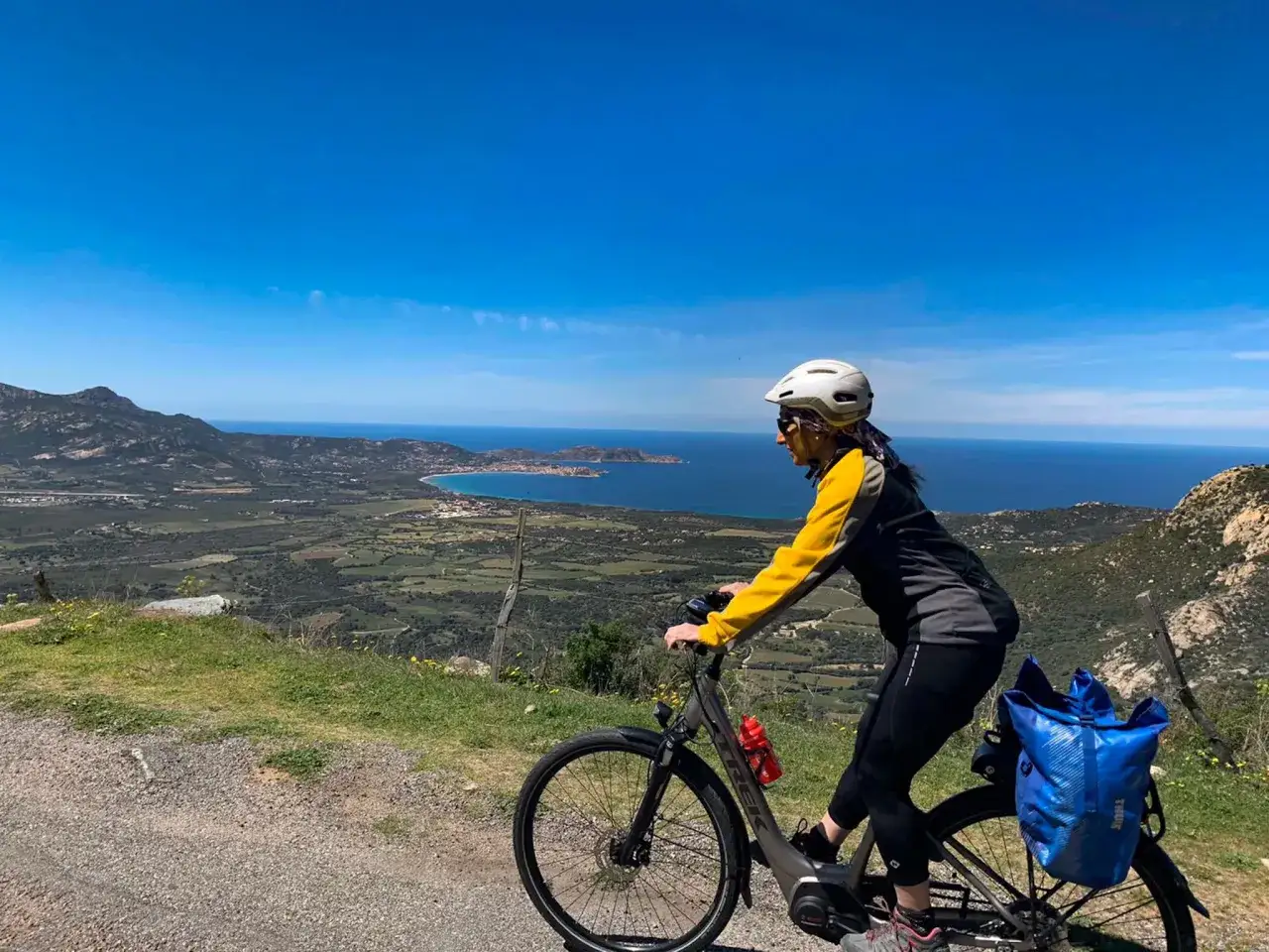 Donna in bici con casco e zaino blu, ammira il panorama mozzafiato della Corsica. Un'avventura indimenticabile in bici.