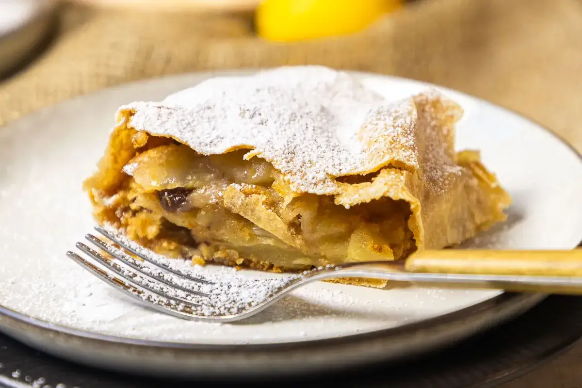 Ein Stück Apfelstrudel, bestäubt mit Puderzucker, liegt auf einem Teller mit einer Gabel.