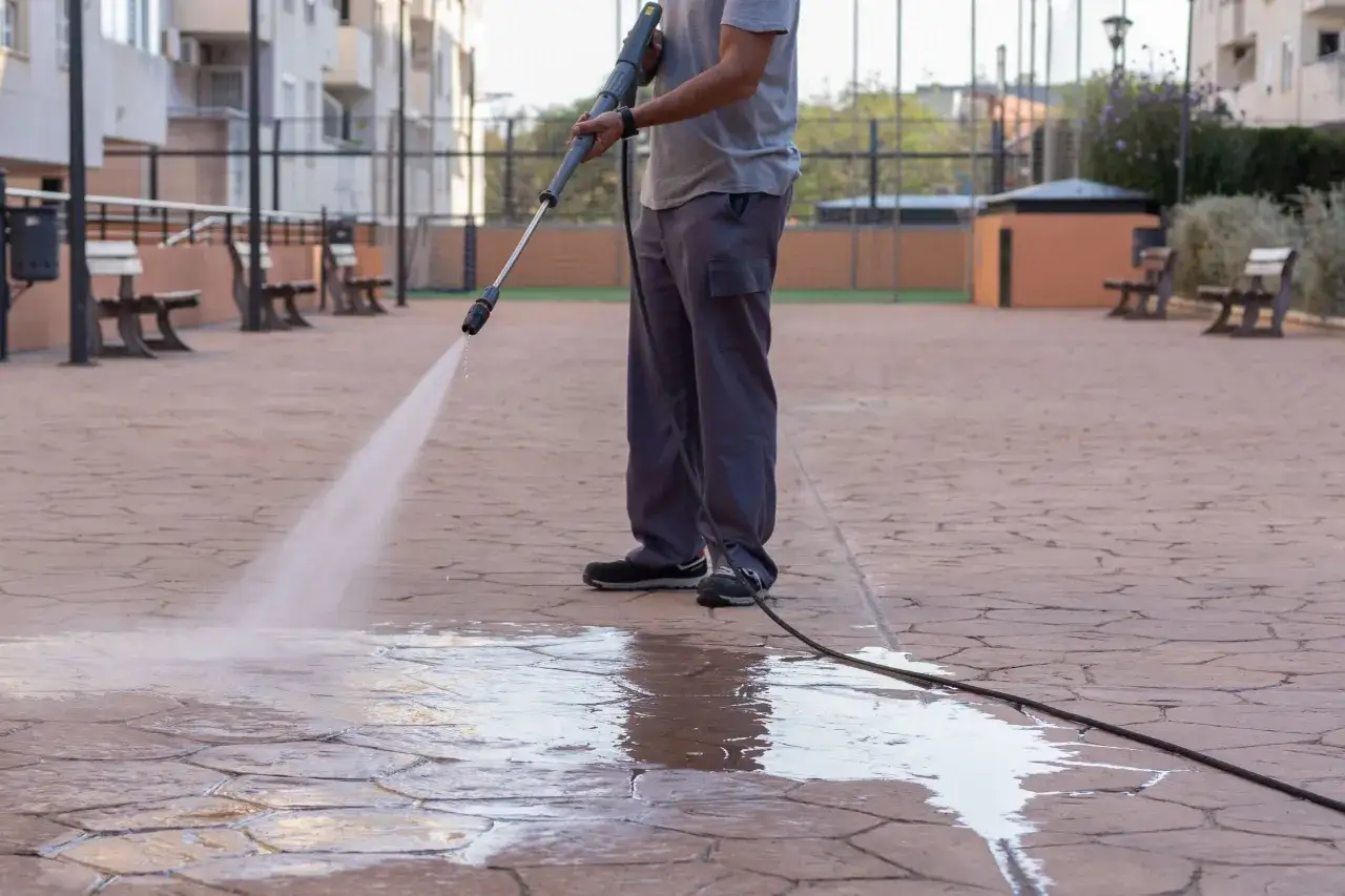 Persona limpiando el suelo de una terraza exterior con una hidrolimpiadora. El agua a presión deja el suelo brillante y limpio.