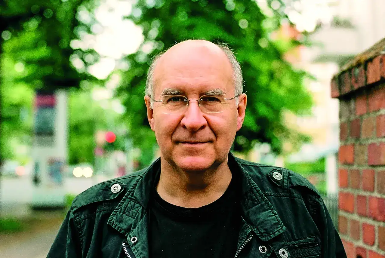 Porträt von Peter Wensierski, einem Mann mit Brille und Glatze, der eine dunkle Jacke trägt. Im Hintergrund sind Bäume und eine Ziegelmauer zu sehen.