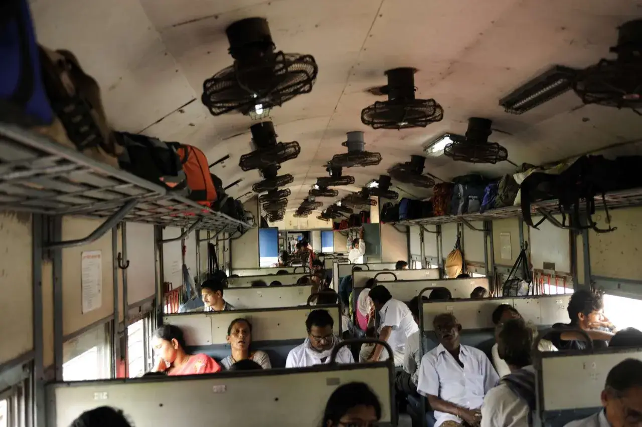 Folk reser i ett fullsatt tåg i Indien, med bagage på hyllorna ovanför och fläktar i taket.