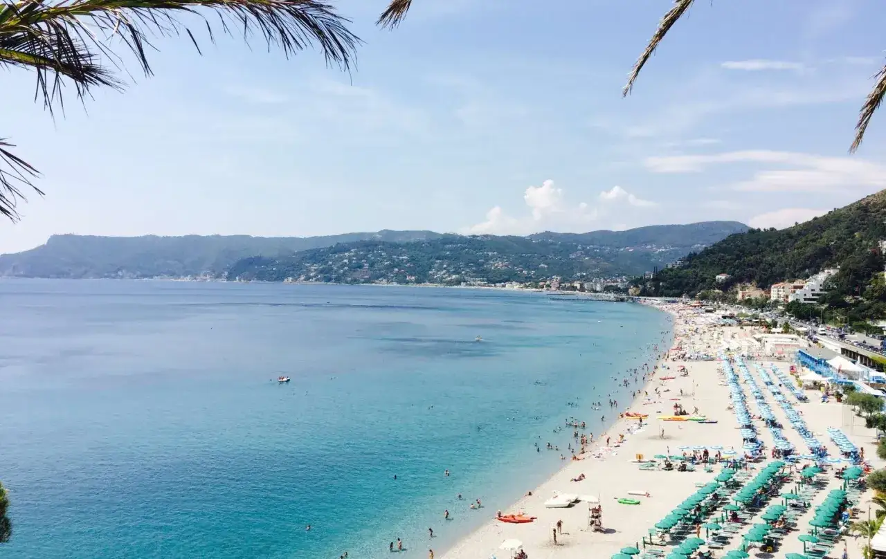 Plage animée sur la Riviera italienne, avec des parasols bleus et verts, des baigneurs dans la mer turquoise et des collines verdoyantes en arrière-plan.