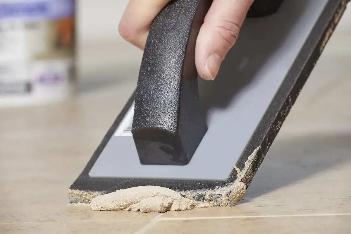 A hand uses a grout float to spread grout between tiles. Wondering how long does it take for grout to set? This image shows the process.