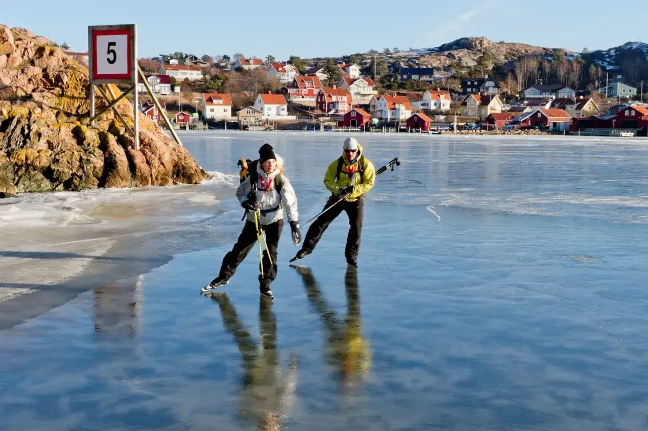 Två skridskoåkare på blank is, med hus och berg i bakgrunden. Frågan är hur tjock ska isen vara för att vara säker?