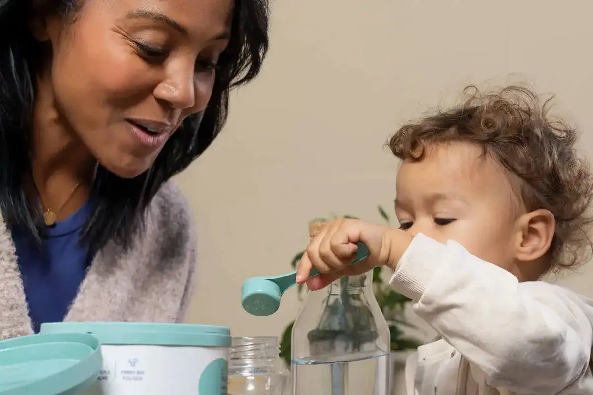 Une mère observe son bébé qui manipule une cuillère doseuse près d'une bouteille d'eau. La question