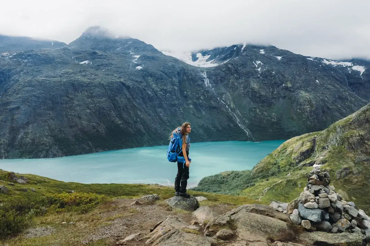 A hiker with a blue backpack stands on a rocky outcrop, overlooking a stunning turquoise lake and majestic mountains in Jotunheimen National Park.