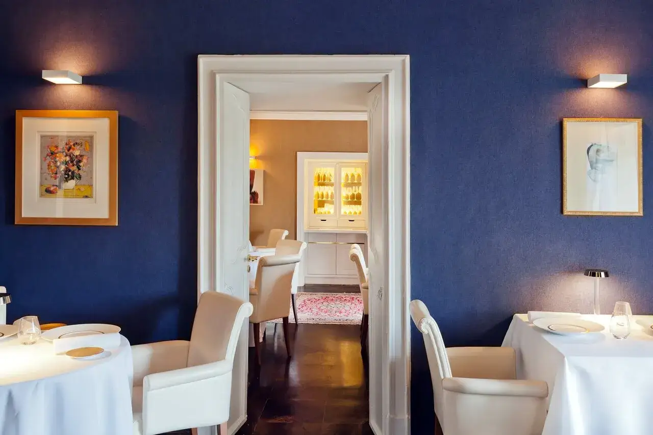 Un ristorante elegante con pareti blu, tavoli apparecchiati e sedie in pelle. Si intravede una sala con un mobile bar illuminato, un'atmosfera da