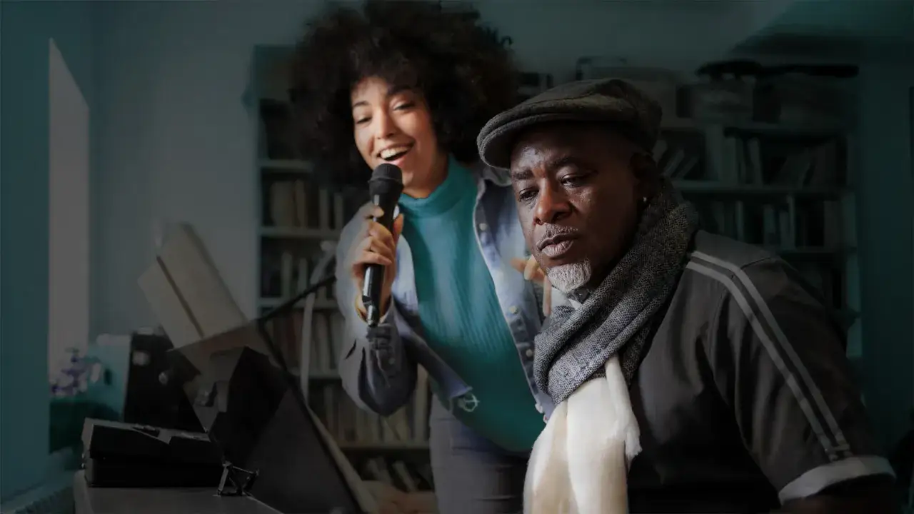 Junge Frau singt ins Mikrofon, w&auml;hrend ein &auml;lterer Mann mit M&uuml;tze und Schal zuh&ouml;rt. Ein Klavier steht im Vordergrund. Der Moment des formant shifting, wenn Musik verbindet.