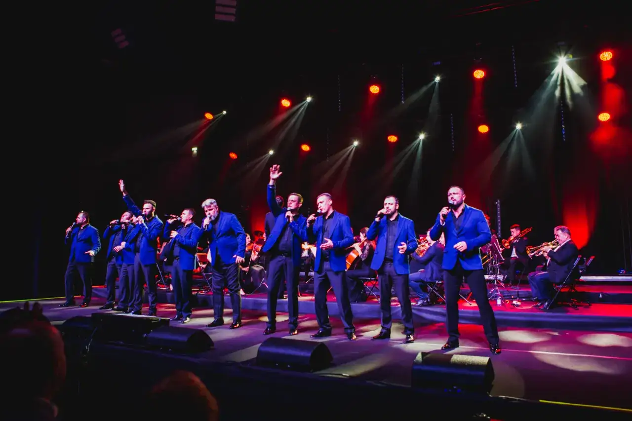 Dziesięciu tenor&oacute;w w niebieskich marynarkach śpiewa na scenie, w tle orkiestra.