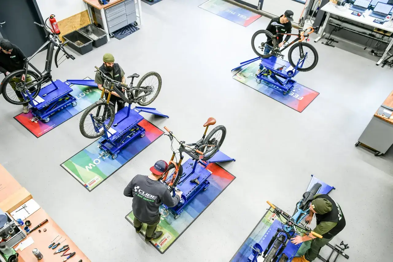 Mehrere Mechaniker führen Fahrrad Wartung in einer Werkstatt durch. Sie arbeiten an verschiedenen Mountainbikes und E-Bikes auf blauen Montageständern.
