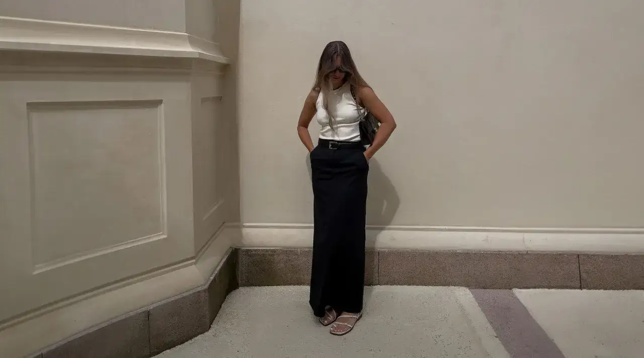 Elegancka stylizacja: biała bluzka do długiej spódnicy, czarna, z paskiem. Kobieta w okularach przeciwsłonecznych i torebce na ramieniu.