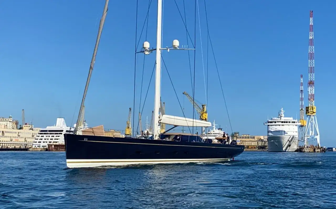 Il kauris iv yacht, scuro e imponente, naviga in un porto con gru e navi da crociera sullo sfondo.