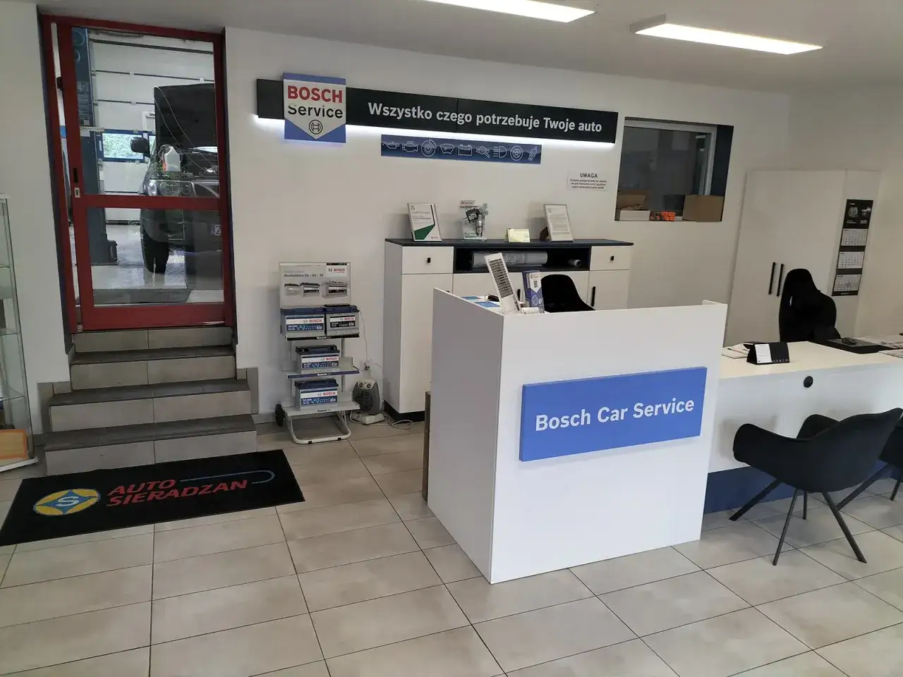 Wnętrze serwisu Bosch Car Service. Na ladzie logo, obok stojaki z akumulatorami. W tle drzwi do warsztatu.