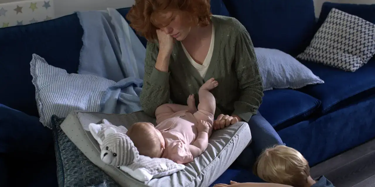 Une mère épuisée, aux prises avec les symptômes du baby blues, s'occupe de son bébé sur un coussin, tandis qu'un autre enfant est allongé à proximité.