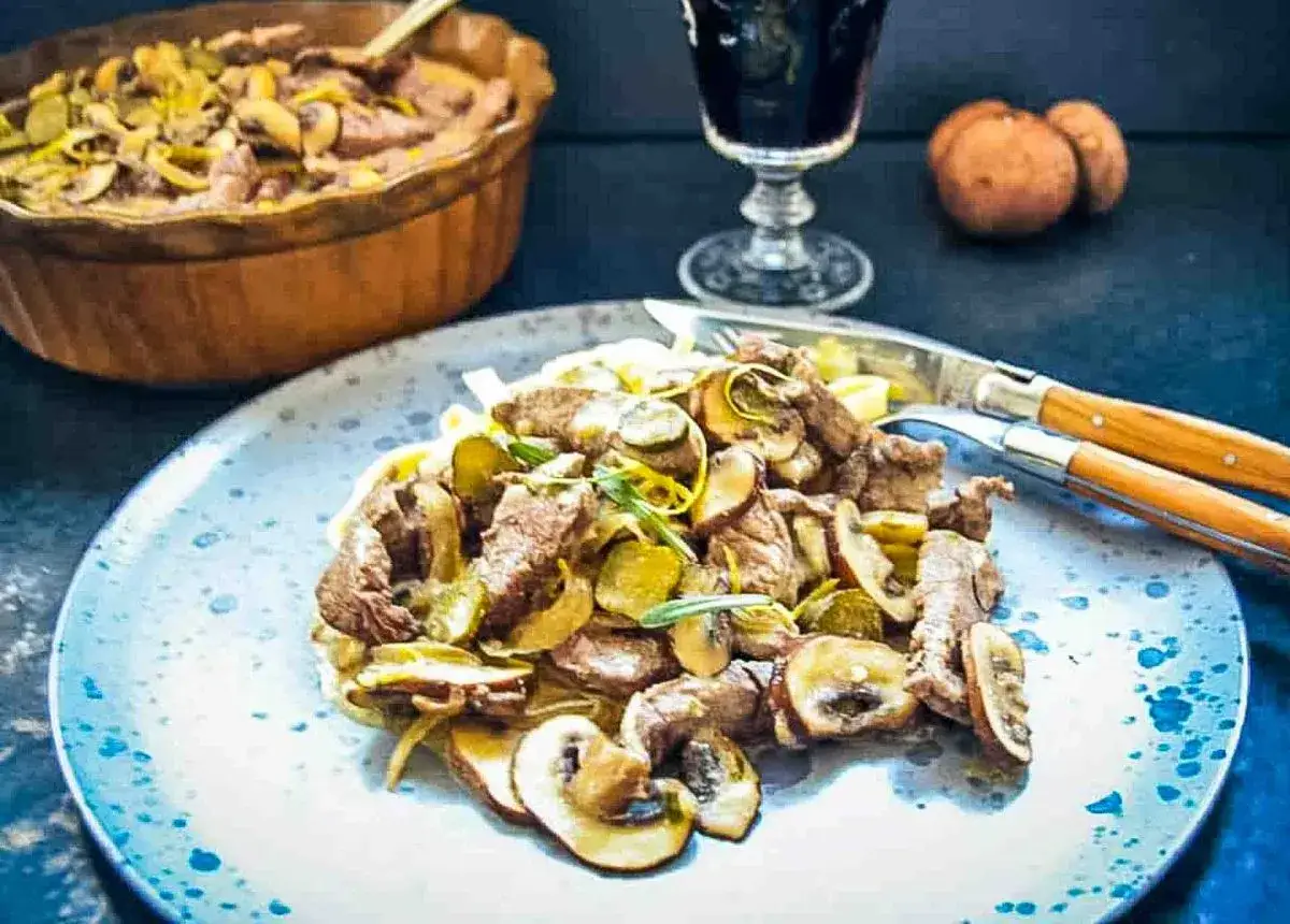 Ein Teller mit einem k&ouml;stlichen Stroganoff Rezept Original, garniert mit Pilzen und Gurken. Daneben ein Glas Rotwein und eine Sch&uuml;ssel mit dem Gericht.