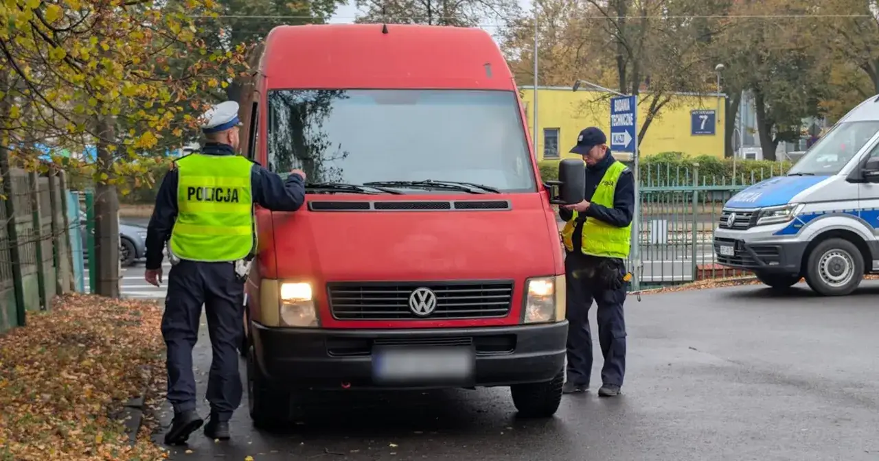 Policjanci kontrolują czerwonego vana. Jeden sprawdza dokumenty, drugi przygląda się pojazdowi. Brak przeglądu może być przyczyną kontroli.