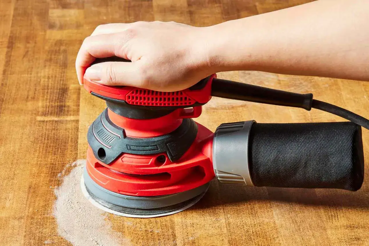 Main d'une personne utilisant une ponceuse de r&eacute;novation rouge sur une surface en bois, cr&eacute;ant de la poussi&egrave;re.