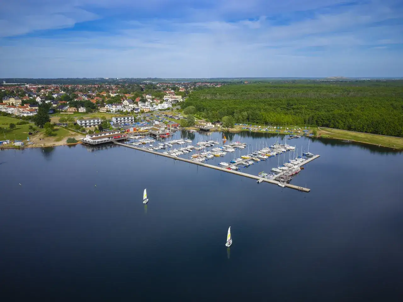 Ein Yachthafen mit vielen Booten und Segelbooten auf dem Wasser, umgeben von einer Stadt und einem Wald. Perfekt f&uuml;r einen Tag an den Badeseen Leipzig.