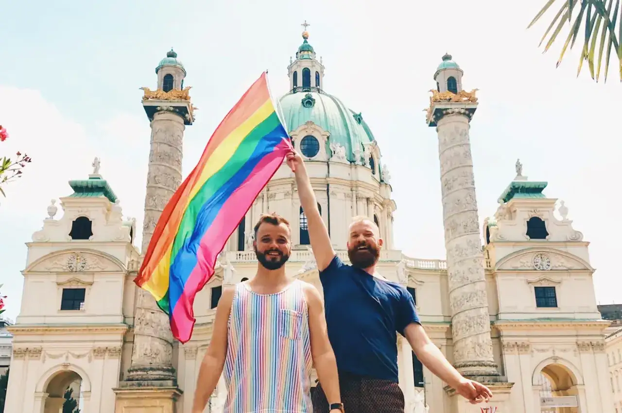 Two men proudly hold a rainbow flag in front of a grand building, celebrating gay Austria.