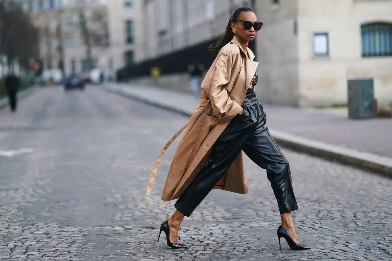 Chic woman in a stylish leather pants outfit for winter, complete with a trench coat and heels, walks down a cobblestone street.