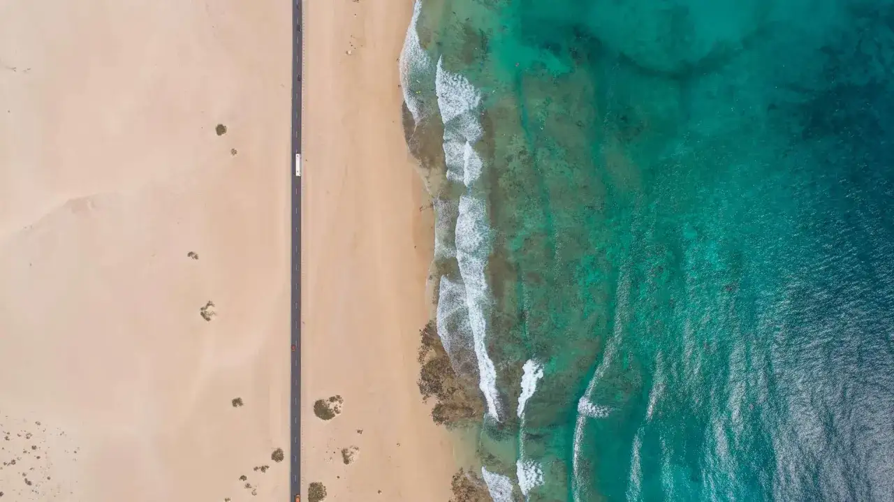Fuerteventura pogoda sierpień: pusta plaża, turkusowe morze i droga wiodąca przez wydmy. Idealna pogoda na wakacje.