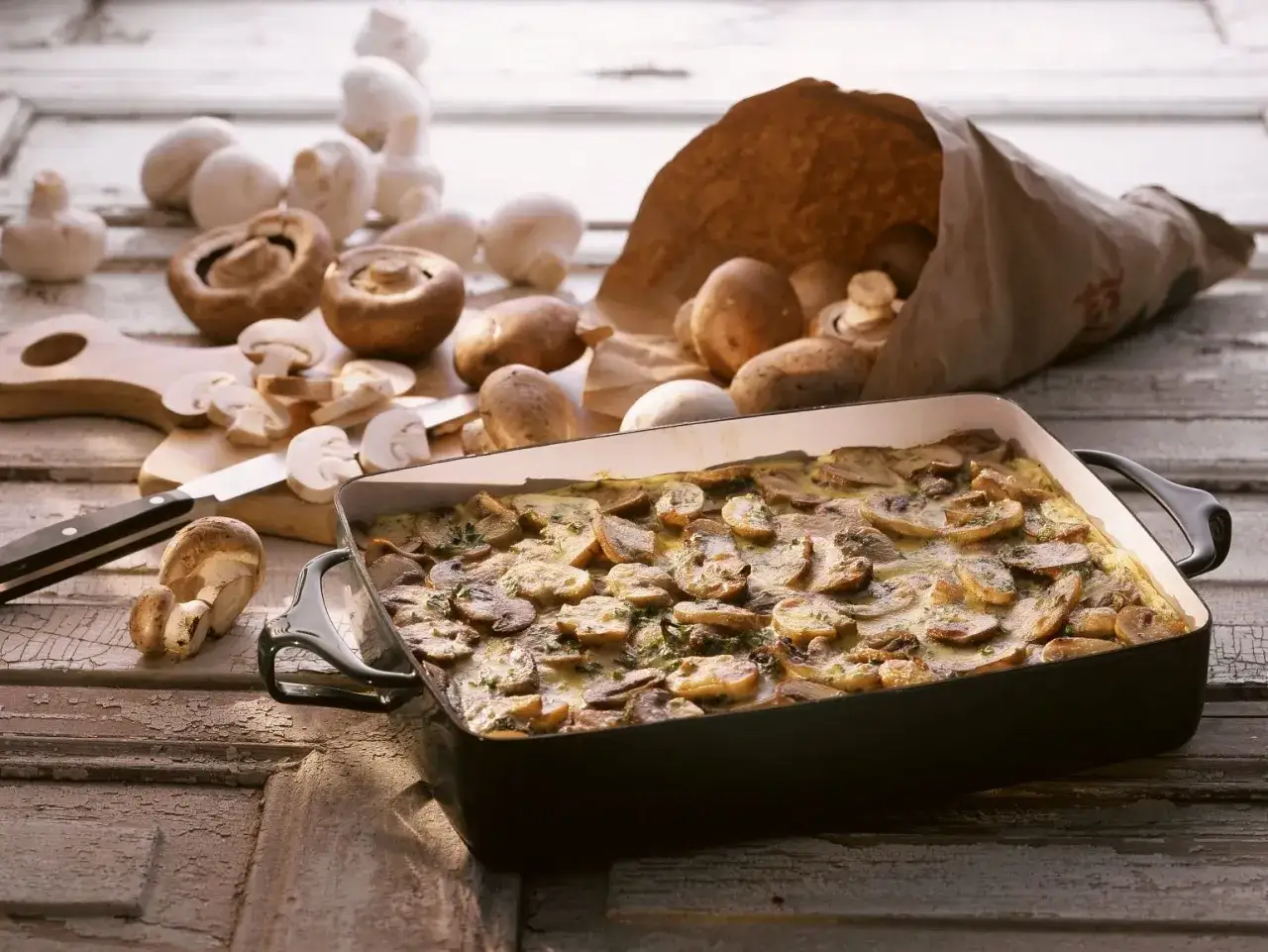 Frisch zubereiteter low carb champignon-auflauf in einer Auflaufform, umgeben von frischen Champignons und einem Messer auf einem Holzbrett.