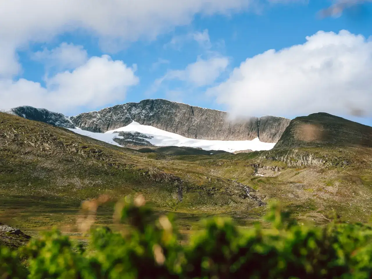 En mäktig helags topptur med snöklädda berg och en blå himmel med moln. Grönskande sluttningar leder upp mot toppen.