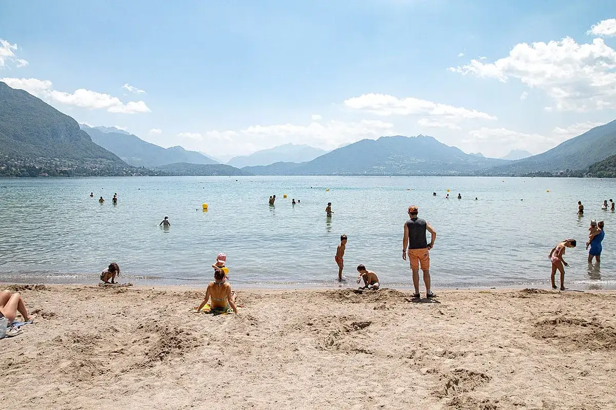 Journée ensoleillée sur la plage d'Annecy. Des familles profitent de l'eau claire et du sable fin, entourées par des montagnes majestueuses.