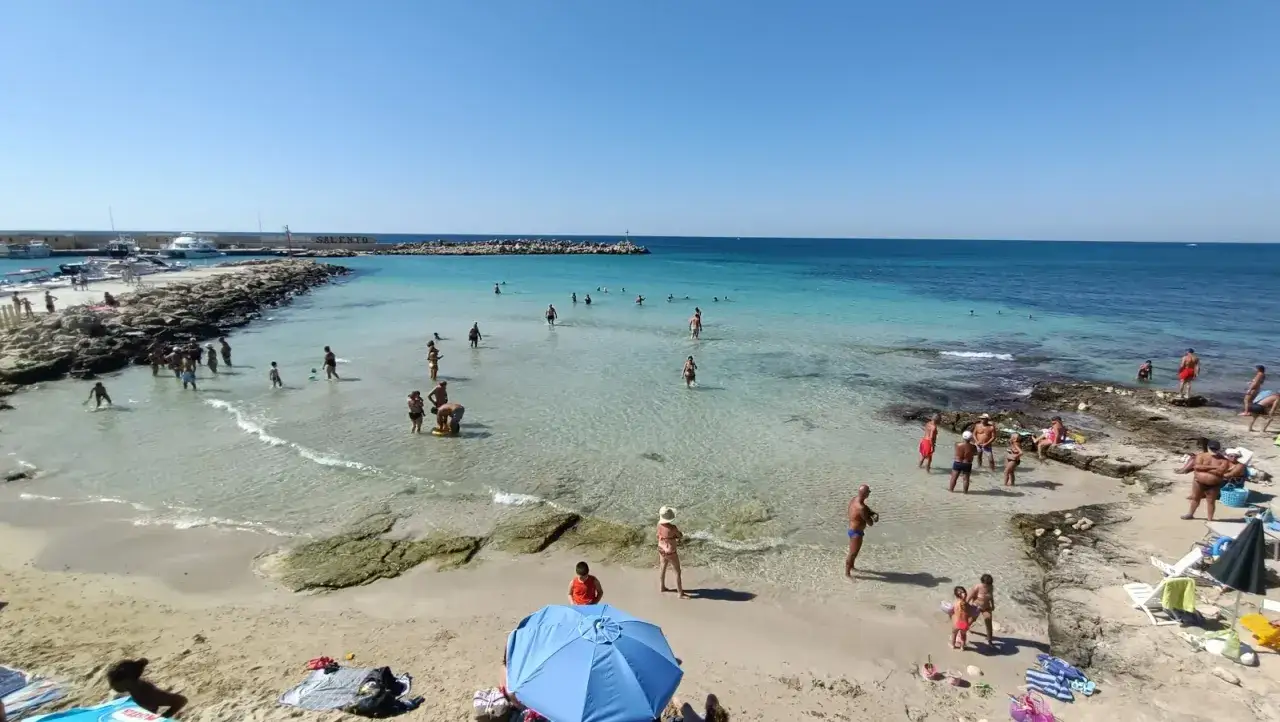Spiaggia affollata a Torre Vado, Lecce, con persone che nuotano e prendono il sole in una giornata limpida.