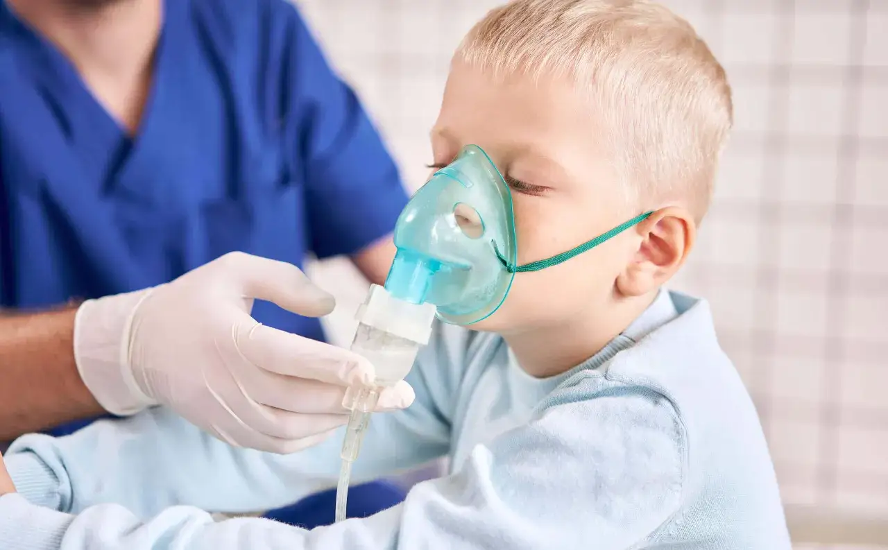 Niño de 3 a 4 años con mascarilla nebulizadora para aliviar tos. Un profesional sanitario con guantes ajusta el dispositivo.