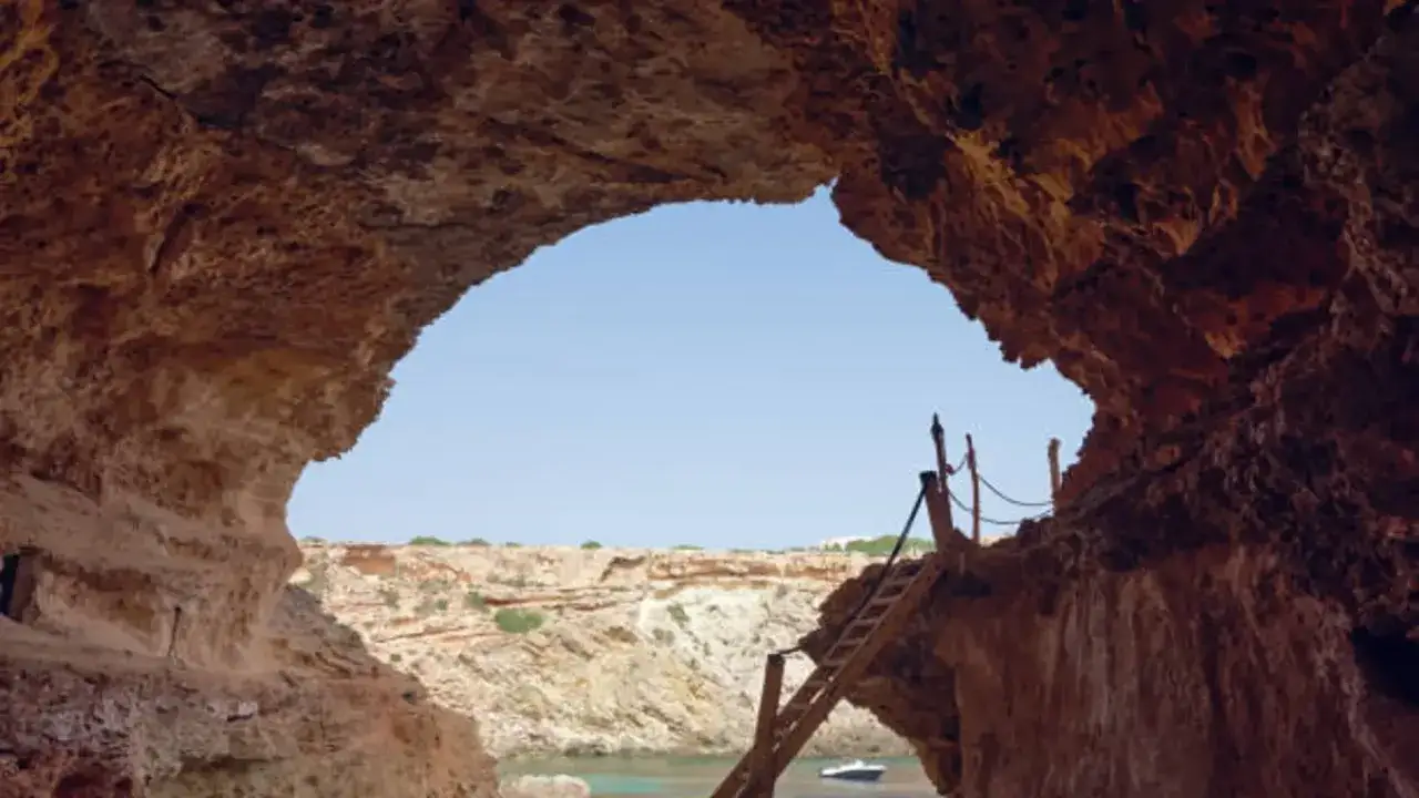 Vista desde una cueva en Cala Comte, con una escalera de madera que desciende hacia el mar azul y un peque&ntilde;o bote.