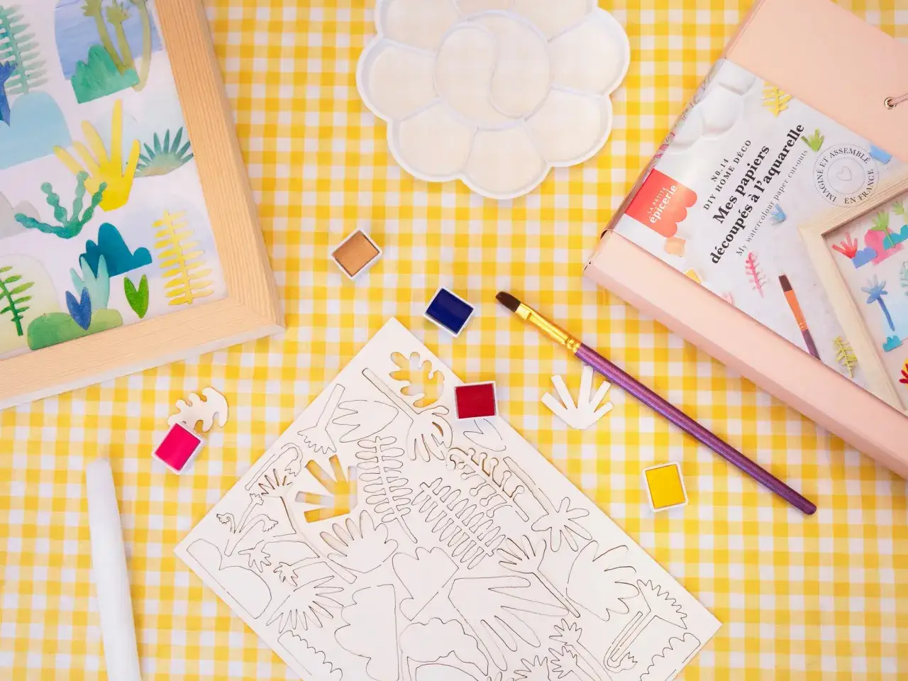 Kit de loisirs cr&eacute;atifs pour un cours d'aquarelle : papiers d&eacute;coup&eacute;s, palette, pinceau et peintures sur une nappe &agrave; carreaux jaunes.