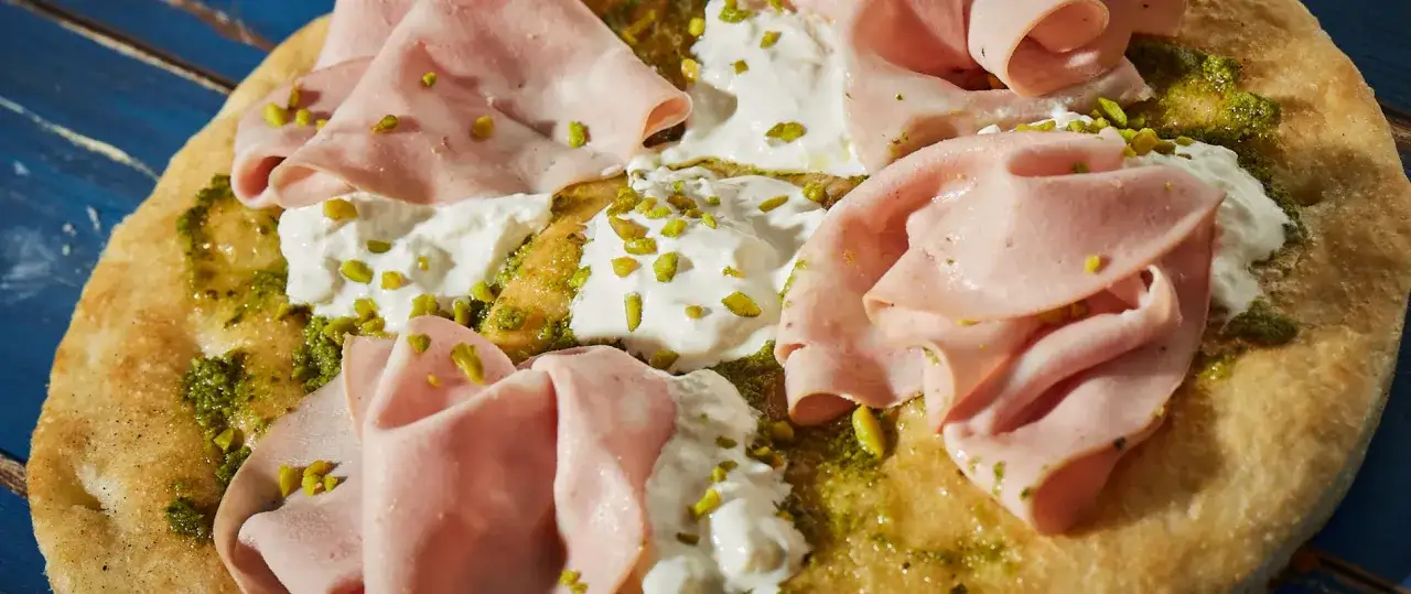 Pizza z mortadelą, kremowym serem, zielonym pesto i pistacjami. Pyszne połączenie smaków na chrupiącym cieście.