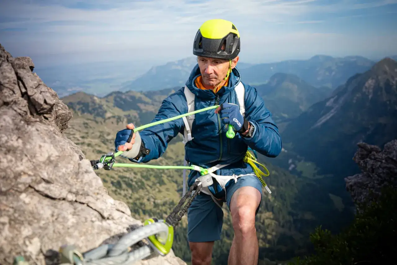 Escalador con casco y arnés, asegurado con un asegurador automático de escalada, en un paisaje montañoso.