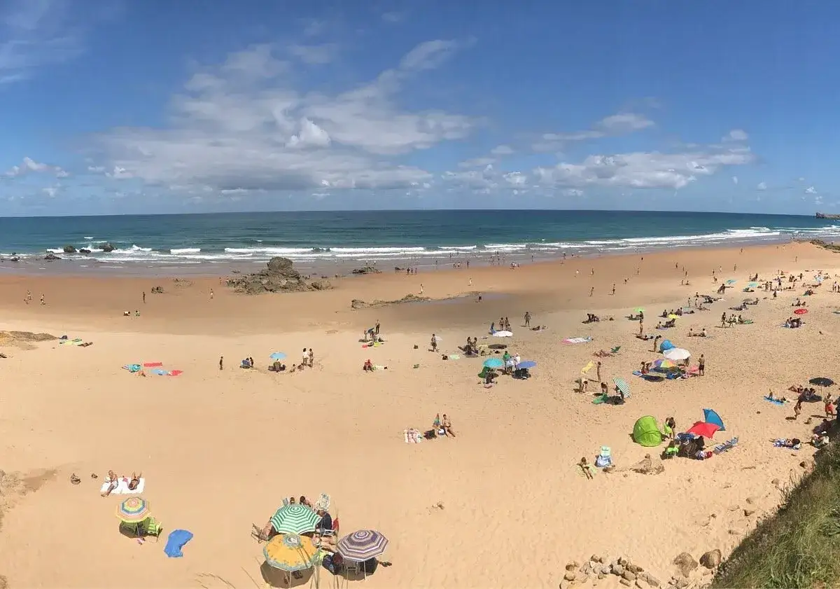 Amplia vista de playas cerca de Comillas con gente disfrutando del sol y el mar bajo un cielo azul con nubes.