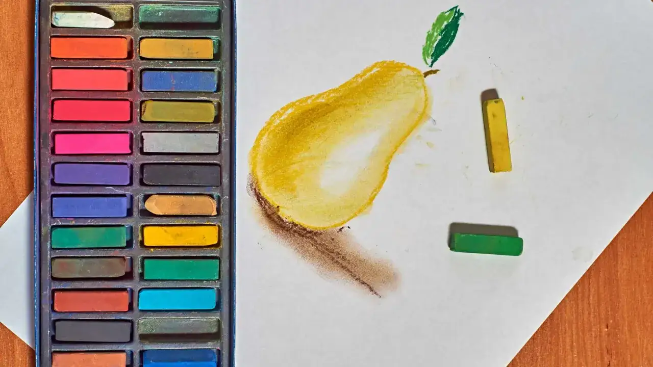 Une poire jaune est dessin&eacute;e &agrave; la **technique pastel sec**. &Agrave; c&ocirc;t&eacute;, une bo&icirc;te de pastels et quelques b&acirc;tonnets de couleur.