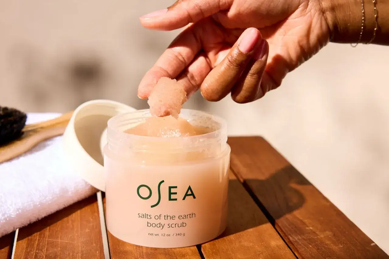 En hand tar en nypa av en rosa kroppsskrubb, en härlig mekanisk peeling för huden. En vit handduk och borste syns i bakgrunden.