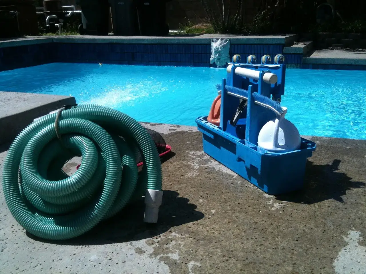 A coiled green hose and a blue pool cleaning caddy sit by a bright blue pool, ready for how to vacuum a pool.