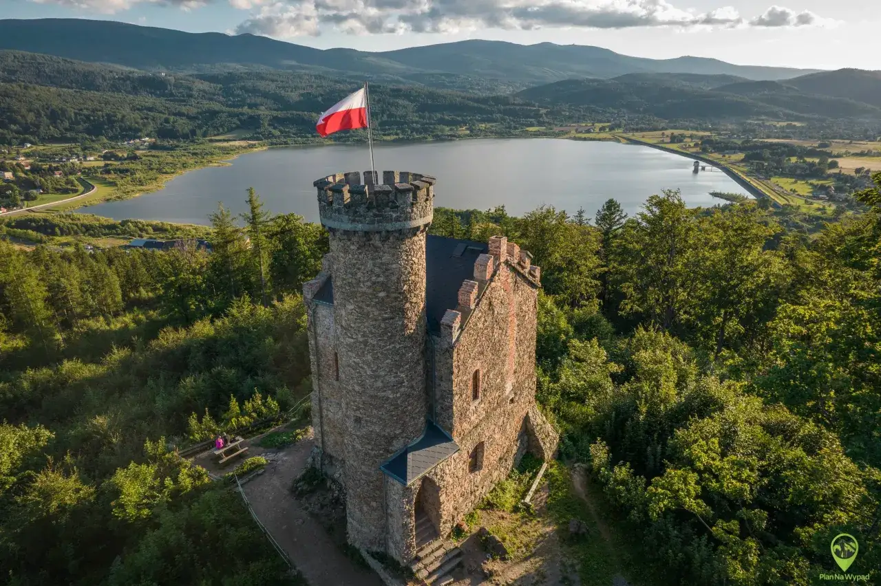 Kamienna wieża widokowa z polską flagą, znana jako zamek księcia Henryka, góruje nad jeziorem i lasami.