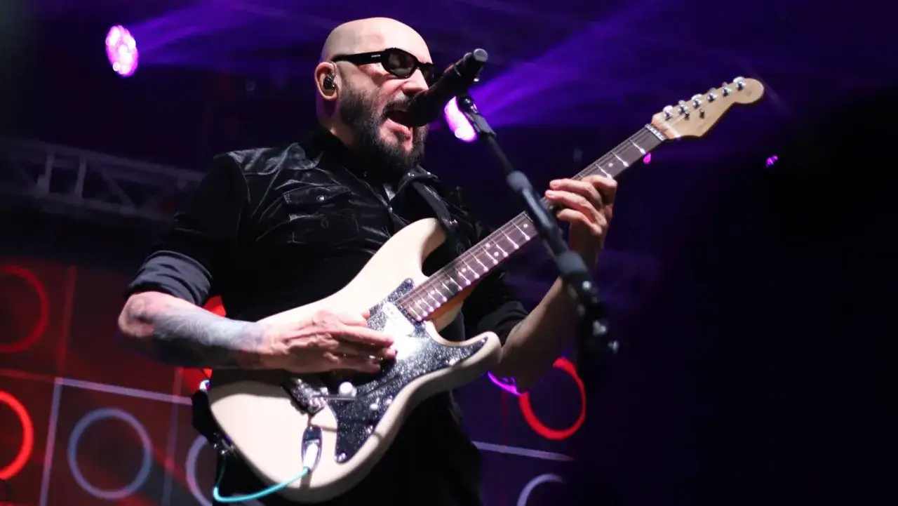 Skawiński w czarnej, aksamitnej koszuli gra na kremowej gitarze elektrycznej, w okularach przeciwsłonecznych, na tle fioletowych świateł.