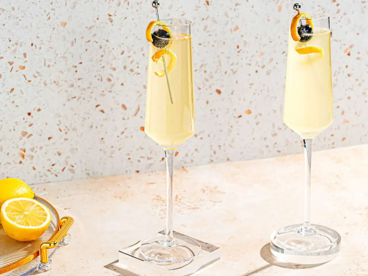 Två eleganta glas med en gyllene French 75, garnerade med citronskal och bär. Citroner ligger på en bricka i förgrunden.