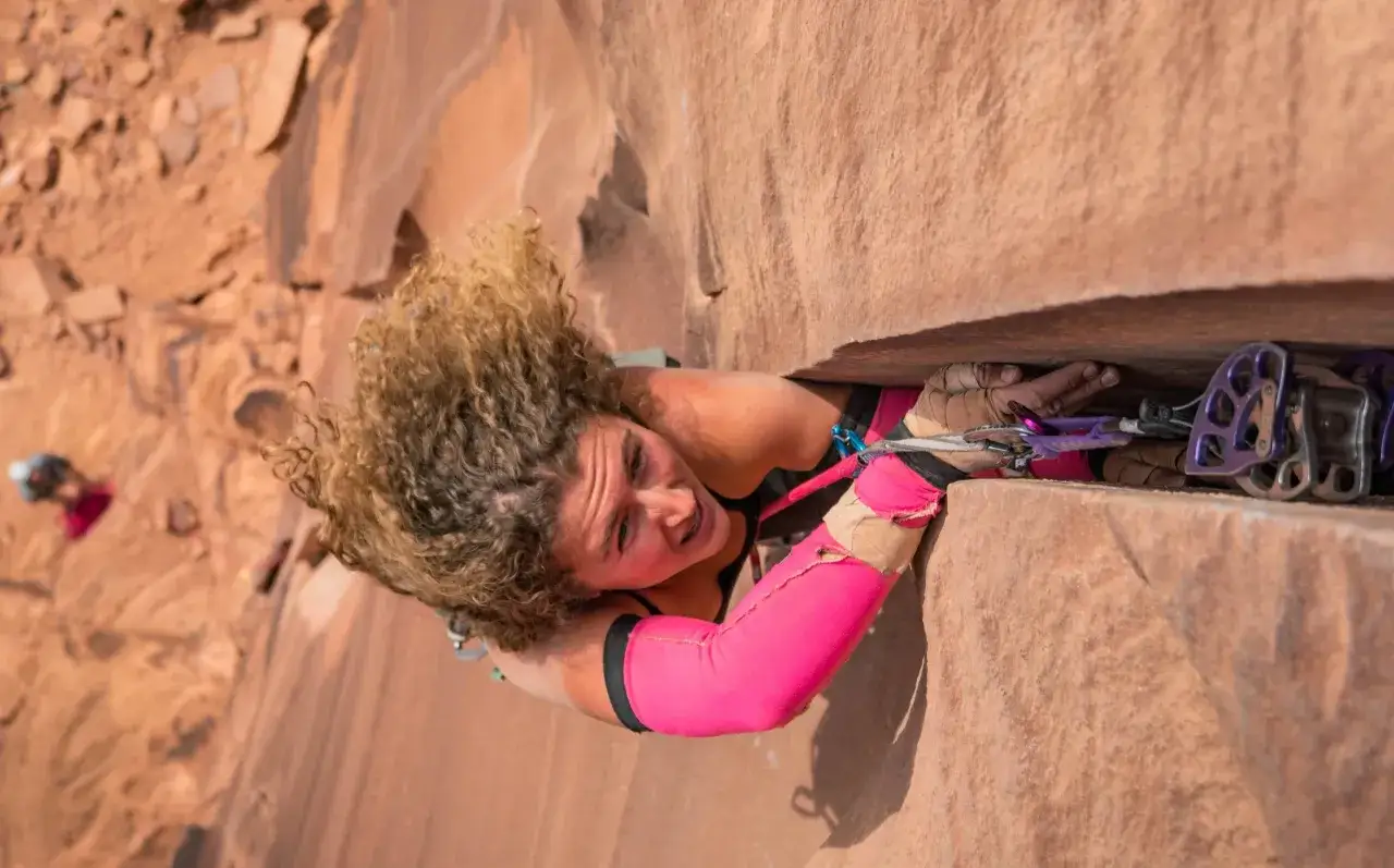 Eine Kletterin meistert eine schwierige Offwidth-Passage in rotem Fels. Ihre lockigen Haare wehen, w&auml;hrend sie sich mit Kraft und Technik nach oben k&auml;mpft.