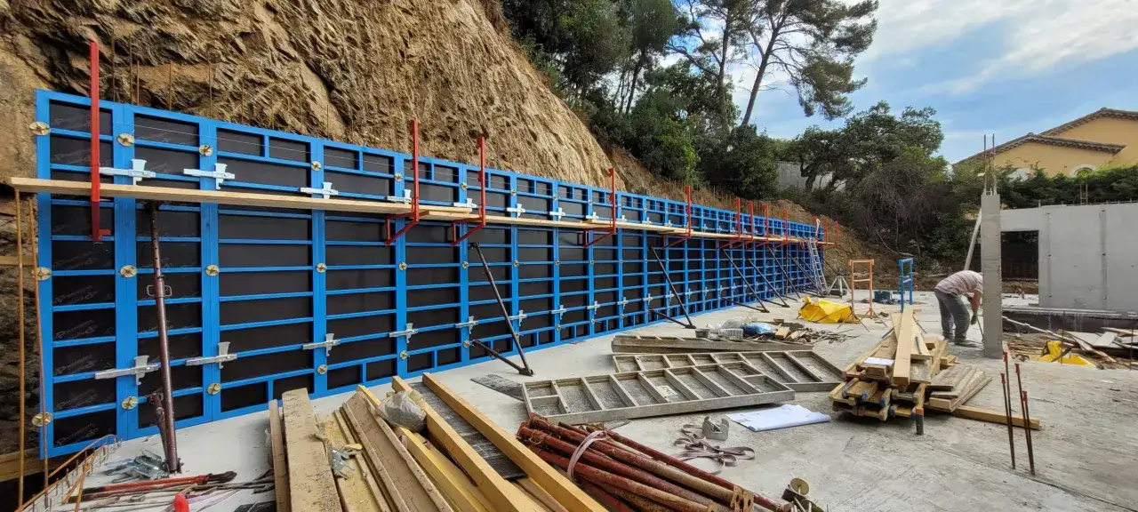 Un mur en béton est en cours de construction. Les ouvriers sont en train de faire un coffrage bleu et noir, soutenu par des étais rouges et métalliques, le long d'une colline rocheuse.
