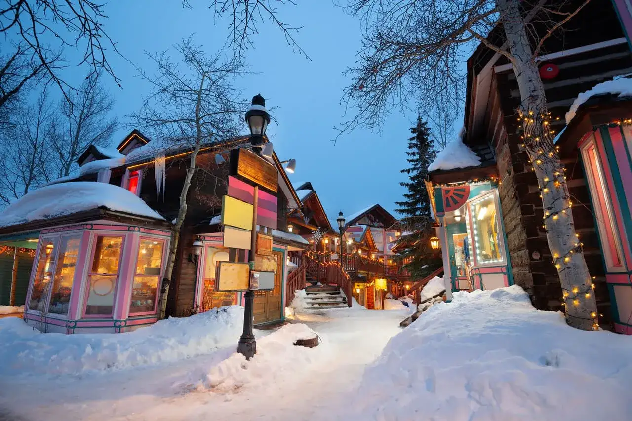 Encantador pueblo nevado, ideal para tus destinos para viajar en enero. Luces cálidas iluminan las tiendas y la nieve cubre el camino.