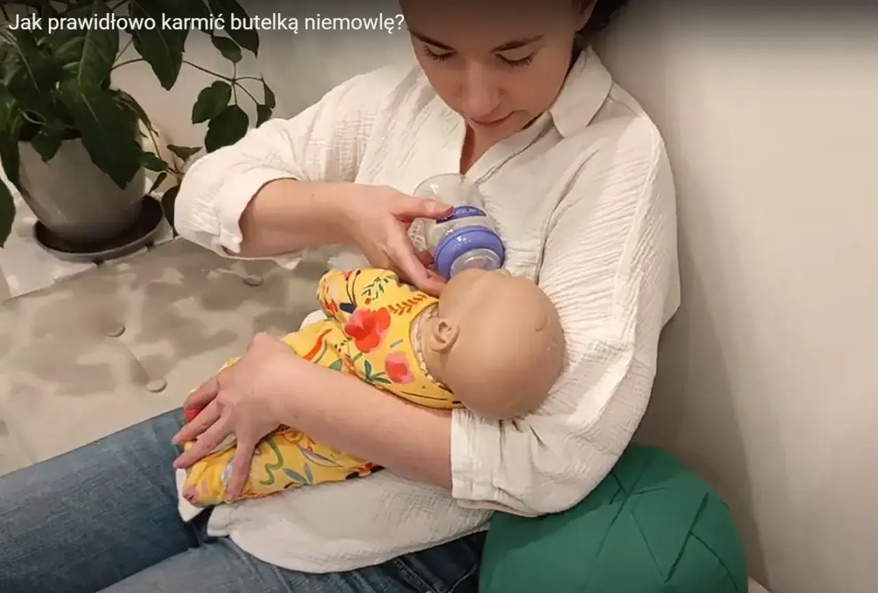 Kobieta uczy się, jak karmić noworodka butelką, trzymając lalkę w ż&oacute;łtym pajacyku w kwiaty.