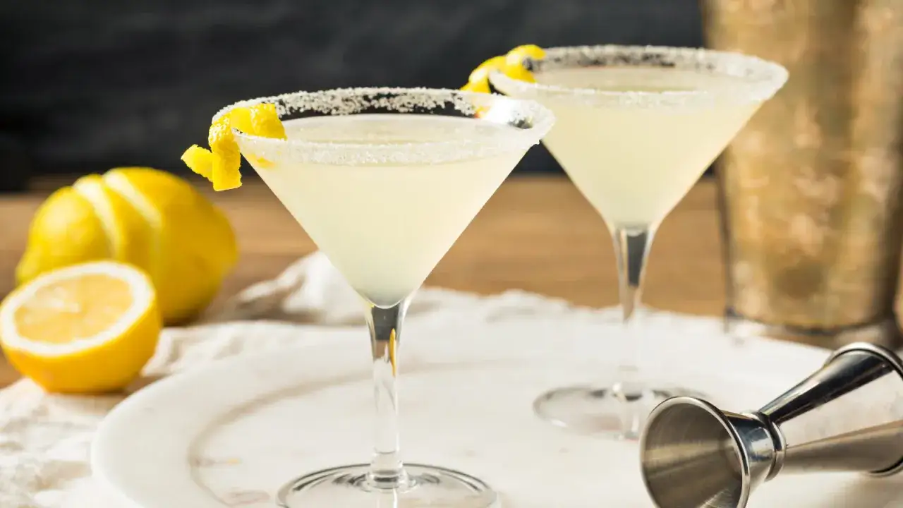 Tv&aring; glas med en ljusgul lemon martini, garnerade med citronskal. Citroner och en shaker i bakgrunden.